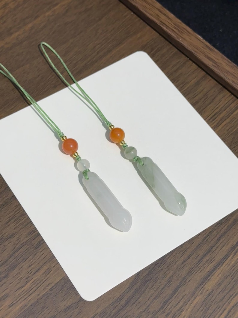 May include: Two decorative charms featuring cylindrical, pale green stone pendants suspended from light green cords. Each charm includes a small, translucent white bead, a vibrant orange bead, and a tiny gold-toned spacer accenting the cord.