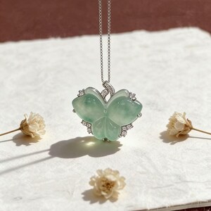 Puede incluir: Un delicado collar de plata con un colgante de jade verde claro con dise&ntilde;o de tr&eacute;bol. El colgante est&aacute; adornado con peque&ntilde;as piedras preciosas transparentes. El collar se exhibe sobre una superficie blanca con flores secas.