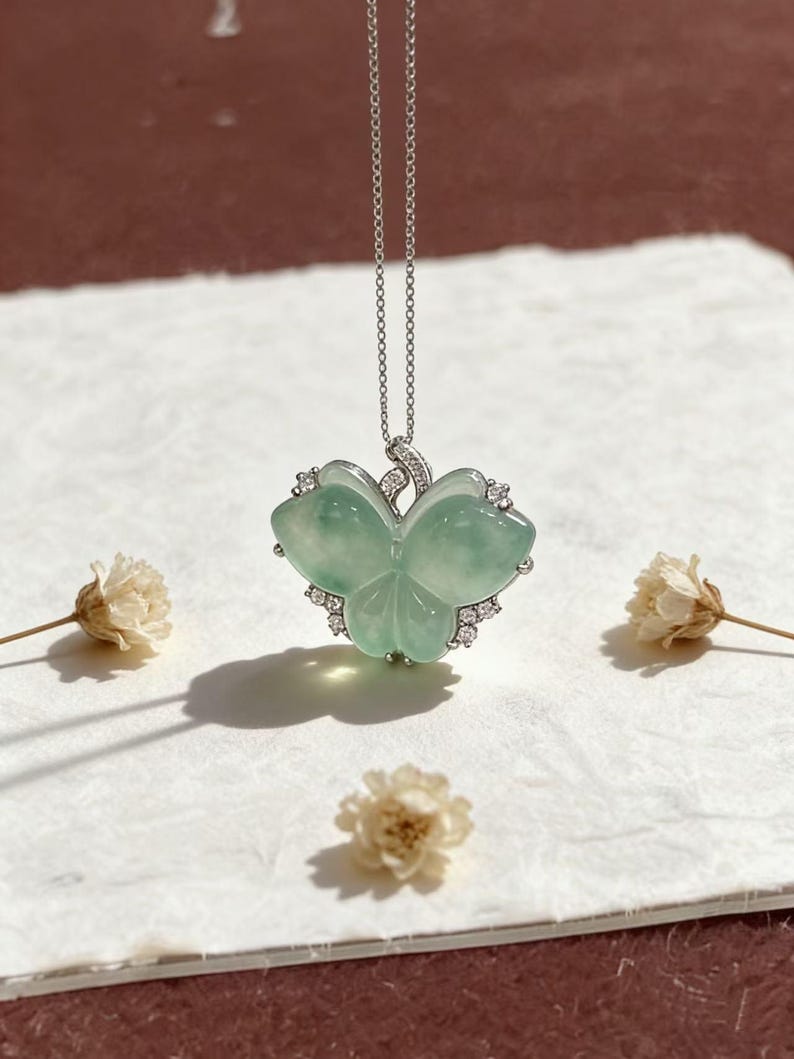 Puede incluir: Un delicado collar de plata con un colgante de jade verde claro con dise&ntilde;o de tr&eacute;bol. El colgante est&aacute; adornado con peque&ntilde;as piedras preciosas transparentes. El collar se exhibe sobre una superficie blanca con flores secas.