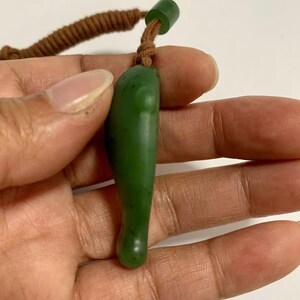 Puede incluir: Un colgante de jade verde con forma de l&aacute;grima, suspendido de un cord&oacute;n marr&oacute;n. El jade tiene una superficie lisa y pulida y un color verde oscuro. El cord&oacute;n est&aacute; anudado en la parte superior y tiene una peque&ntilde;a cuenta verde.