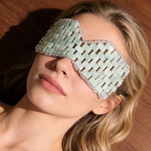 Puede incluir: Una m&aacute;scara de ojos de jade verde claro con un dise&ntilde;o tejido. La m&aacute;scara est&aacute; bordeada con peque&ntilde;as cuentas transparentes. El fondo es una superficie de madera. La m&aacute;scara est&aacute; dise&ntilde;ada para usarse sobre los ojos.