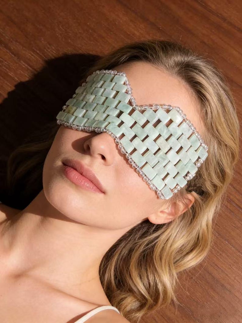 Puede incluir: Una m&aacute;scara de ojos de jade verde claro con un dise&ntilde;o tejido. La m&aacute;scara est&aacute; bordeada con peque&ntilde;as cuentas transparentes. El fondo es una superficie de madera. La m&aacute;scara est&aacute; dise&ntilde;ada para usarse sobre los ojos.