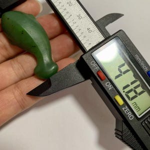 Puede incluir: Una talla en forma de delf&iacute;n, de color verde oscuro, se mide con un calibrador digital. El calibrador muestra una medida de 0,418 cm. La talla se sostiene en una mano y el calibrador es negro.