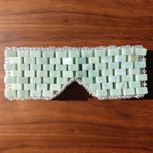 Puede incluir: Una m&aacute;scara para los ojos de jade verde claro con un patr&oacute;n de ladrillos. La m&aacute;scara est&aacute; bordeada con cuentas transparentes. La m&aacute;scara est&aacute; dise&ntilde;ada para cubrir los ojos y est&aacute; sobre una superficie de madera.