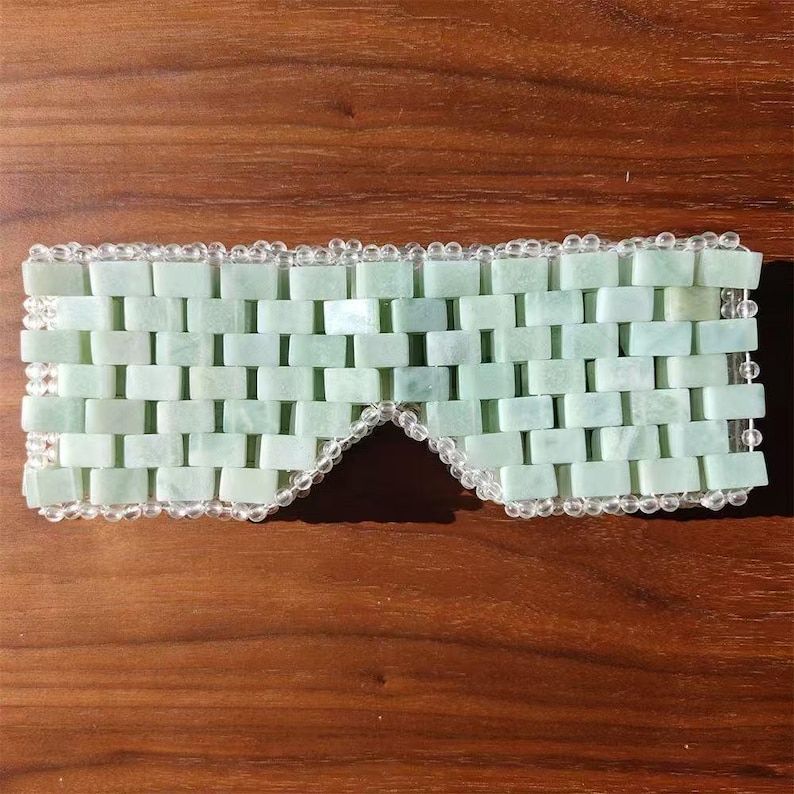 Puede incluir: Una m&aacute;scara para los ojos de jade verde claro con un patr&oacute;n de ladrillos. La m&aacute;scara est&aacute; bordeada con cuentas transparentes. La m&aacute;scara est&aacute; dise&ntilde;ada para cubrir los ojos y est&aacute; sobre una superficie de madera.