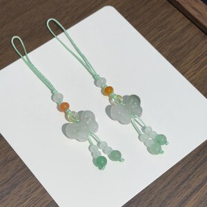 May include: Two decorative charms featuring carved light green stone butterflies. Each charm is attached to a mint green cord and accented with small round beads in shades of white, orange, and green, ending in two dangling beaded strands.