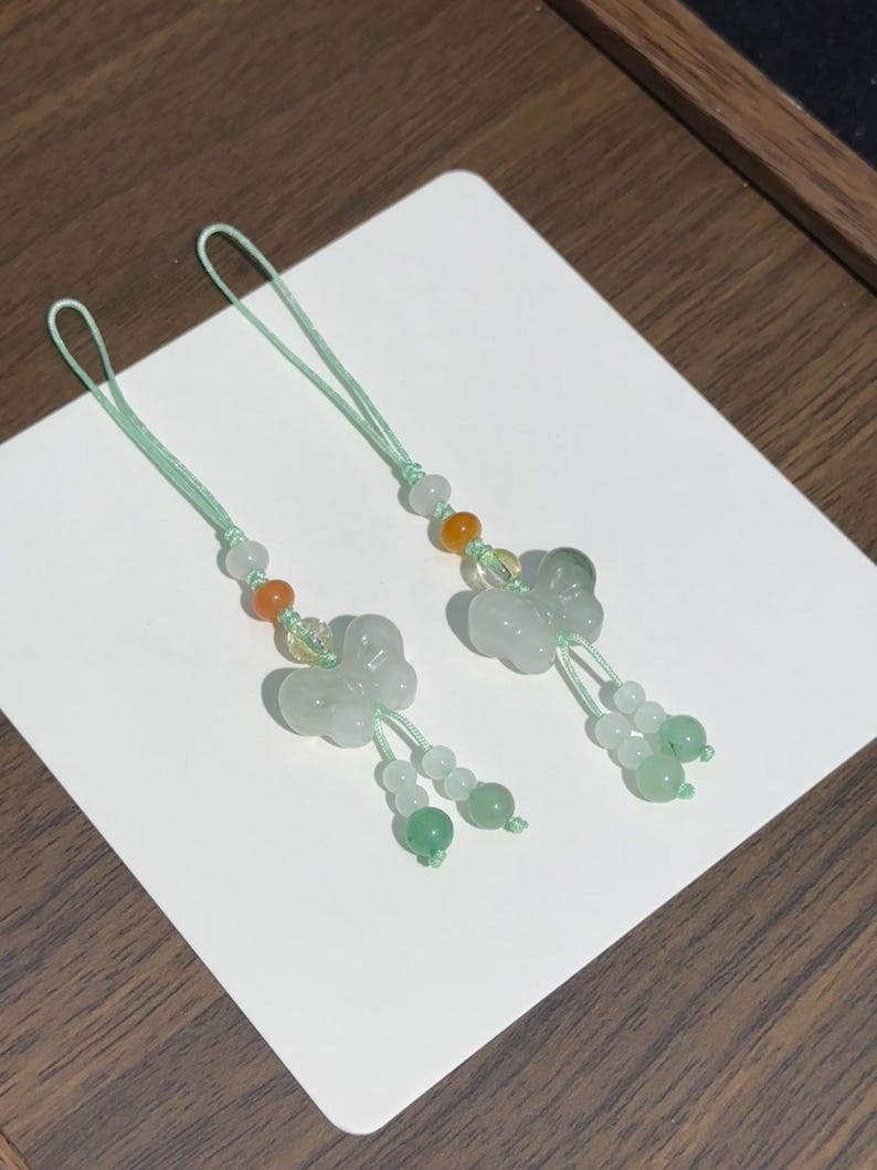 May include: Two decorative charms featuring carved light green stone butterflies. Each charm is attached to a mint green cord and accented with small round beads in shades of white, orange, and green, ending in two dangling beaded strands.
