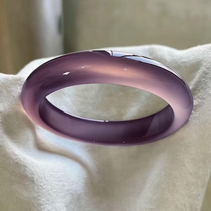 May include: A translucent, lavender-colored bangle bracelet. The smooth, polished surface reflects light, showcasing a gradient of purple hues. The bracelet is a circular shape, likely worn on the wrist. The jewelry is set against a white fabric background.