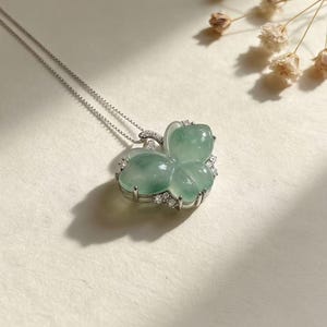 Puede incluir: Un collar de plata con un colgante de mariposa. El colgante est&aacute; hecho de una piedra preciosa verde p&aacute;lido, con forma de mariposa, y adornado con peque&ntilde;as piedras preciosas transparentes. El collar se muestra sobre una superficie de color claro.