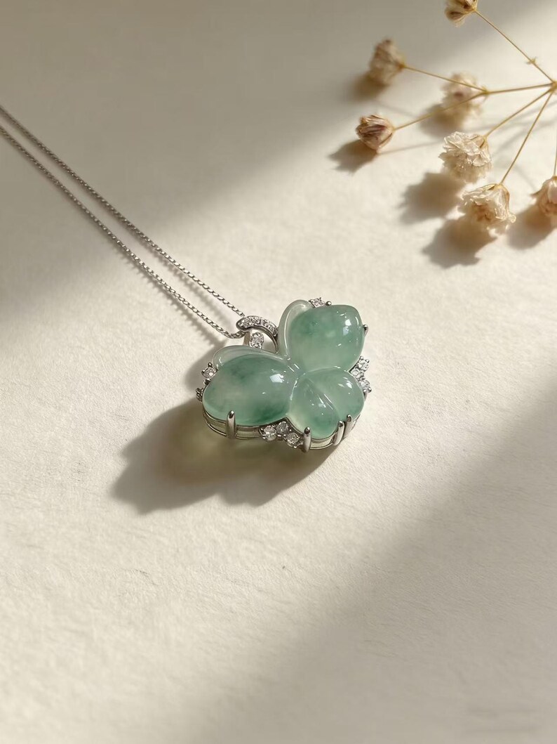 Puede incluir: Un collar de plata con un colgante de mariposa. El colgante est&aacute; hecho de una piedra preciosa verde p&aacute;lido, con forma de mariposa, y adornado con peque&ntilde;as piedras preciosas transparentes. El collar se muestra sobre una superficie de color claro.