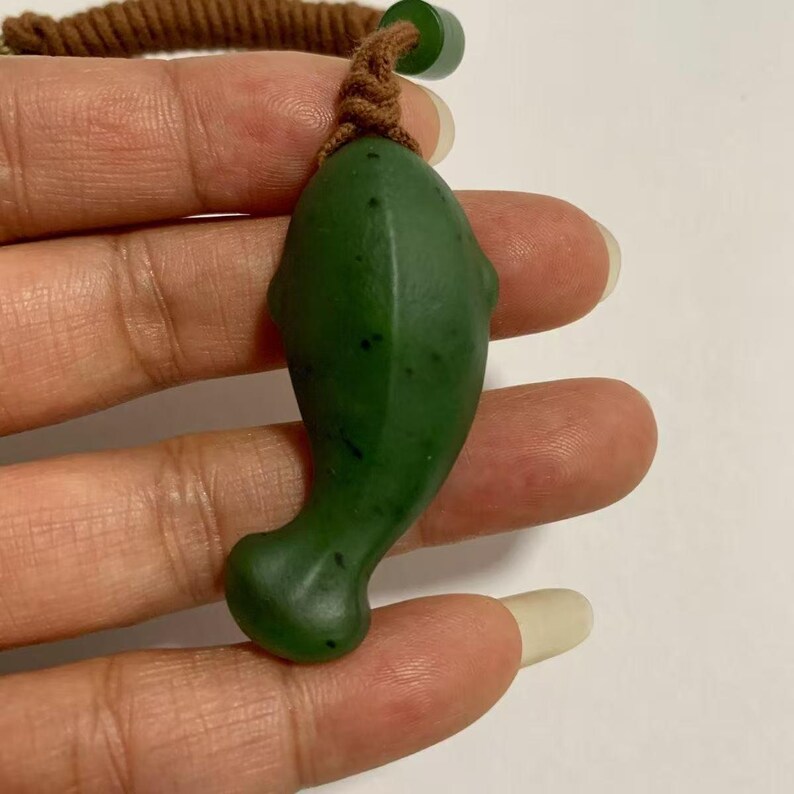 May include: A green jade dolphin pendant with a brown cord. The dolphin-shaped pendant has a smooth, polished surface and a small loop for the cord. The cord is knotted at the top and has a small green bead.