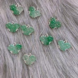 Puede incluir: Una colecci&oacute;n de collares con colgantes de mariposa. Cada colgante presenta un dise&ntilde;o de mariposa verde transl&uacute;cida con detalles blancos y peque&ntilde;as gemas transparentes. Los collares se exhiben sobre una superficie gris texturizada.