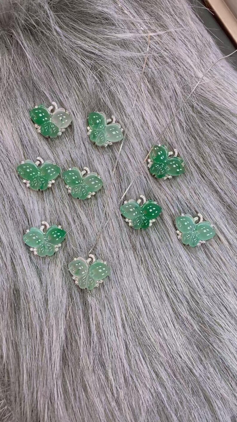 Puede incluir: Una colecci&oacute;n de collares con colgantes de mariposa. Cada colgante presenta un dise&ntilde;o de mariposa verde transl&uacute;cida con detalles blancos y peque&ntilde;as gemas transparentes. Los collares se exhiben sobre una superficie gris texturizada.