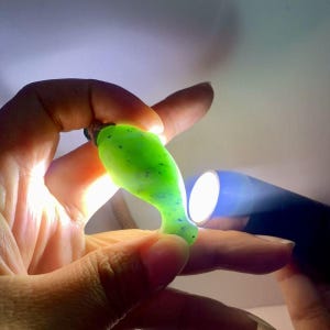 Puede incluir: Un colgante verde brillante en forma de l&aacute;grima con motas azul oscuro. El colgante se sostiene en la mano de una persona e iluminado por una linterna. El colgante tiene una superficie lisa y pulida y un peque&ntilde;o lazo en la parte superior para un collar.