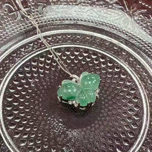 Puede incluir: Un collar de plata con un colgante en forma de mariposa. El colgante est&aacute; hecho de jade verde y est&aacute; adornado con peque&ntilde;as piedras preciosas transparentes. El collar se muestra sobre un plato de vidrio transparente texturizado.