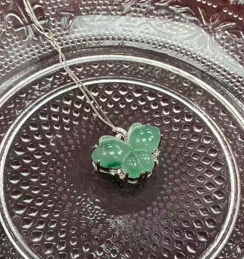 Puede incluir: Un collar de plata con un colgante en forma de mariposa. El colgante est&aacute; hecho de jade verde y est&aacute; adornado con peque&ntilde;as piedras preciosas transparentes. El collar se muestra sobre un plato de vidrio transparente texturizado.