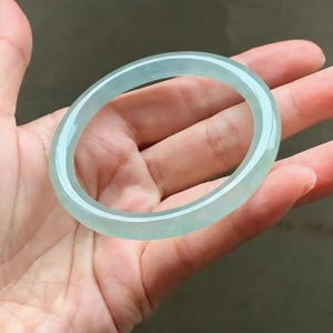 May include: A translucent, pale green jadeite bangle bracelet. The smooth, polished surface reflects light, showcasing the natural variations in color. The circular design is a classic jewelry style.
