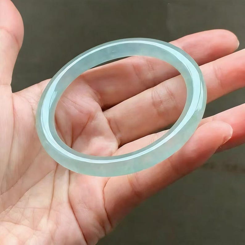 May include: A translucent, pale green jadeite bangle bracelet. The smooth, polished surface reflects light, showcasing the natural variations in color. The circular design is a classic jewelry style.