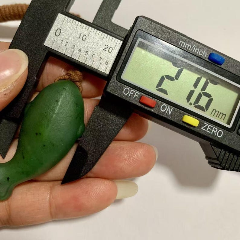 Puede incluir: Un colgante verde con forma de delf&iacute;n se mide con un calibrador digital. El calibrador muestra una medida de 21,6 mm. El colgante est&aacute; sujeto por una mano.