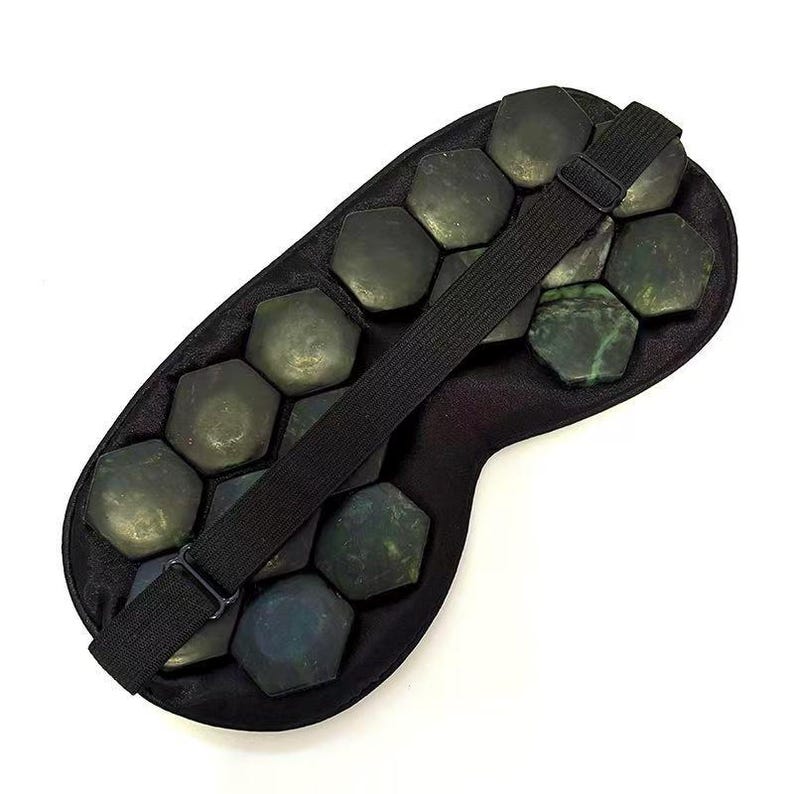 May include: A black eye mask filled with dark green, hexagonal stones. A black elastic strap is attached to the mask, designed to secure it around the head. The stones appear to be jade, and the mask is designed for relaxation.