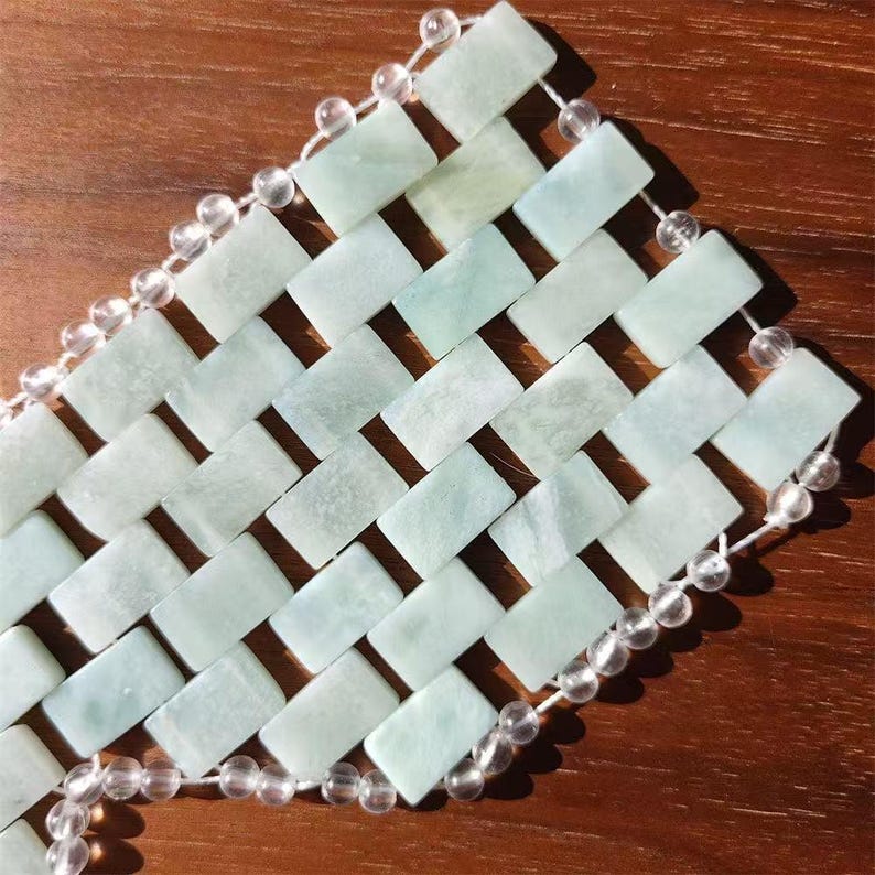 Puede incluir: Una pulsera de jade tejida con piedras rectangulares en tonos verde p&aacute;lido y blanco. Los bordes est&aacute;n adornados con cuentas redondas y transparentes ensartadas en un cord&oacute;n blanco. La pulsera se muestra sobre una superficie de madera.
