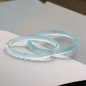 May include: Two light blue jade bangles, each with a smooth, polished surface. The bangles are circular and appear translucent, resting on a white surface. The overall aesthetic is minimalist and elegant, suggesting a focus on natural materials and simple design.