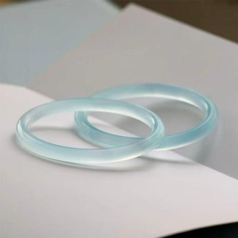 May include: Two light blue jade bangles, each with a smooth, polished surface. The bangles are circular and appear translucent, resting on a white surface. The overall aesthetic is minimalist and elegant, suggesting a focus on natural materials and simple design.