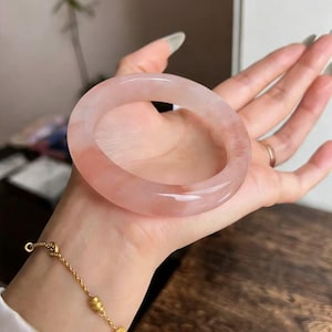 May include: A translucent, pink gemstone bracelet. The bracelet is a solid band of polished stone, showcasing variations in color. A delicate gold bracelet with small charms is also visible.