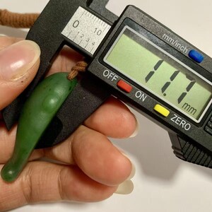 Puede incluir: Un colgante de jade verde, medido en 11,7 mm, se sostiene junto a un calibrador digital. El colgante tiene una forma lisa y alargada con un peque&ntilde;o lazo para un cord&oacute;n. El calibrador muestra la medida en mil&iacute;metros.