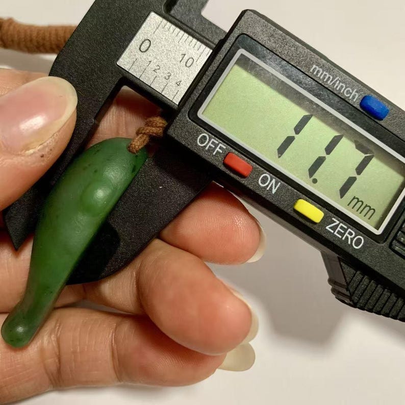 Puede incluir: Un colgante de jade verde, medido en 11,7 mm, se sostiene junto a un calibrador digital. El colgante tiene una forma lisa y alargada con un peque&ntilde;o lazo para un cord&oacute;n. El calibrador muestra la medida en mil&iacute;metros.