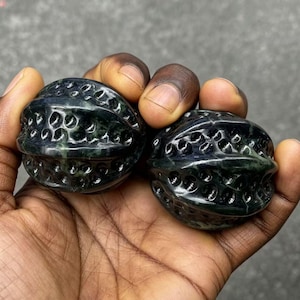 Pode incluir: Duas esferas de pedra esculpida verde escuro com um design vazado texturizado. As esferas são seguradas na mão de uma pessoa, mostrando os detalhes intrincados e a superfície polida da pedra. As esferas têm uma forma orgânica única.