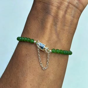 May include: A green beaded bracelet with a silver magnetic clasp and a small chain. The bracelet is made of small, round, green beads. The clasp is a silver cylinder with a small chain attached. The bracelet is on a wrist.