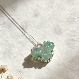 Puede incluir: Un collar de plata con un colgante en forma de mariposa. El colgante est&aacute; hecho de jade verde transl&uacute;cido y est&aacute; adornado con peque&ntilde;as piedras preciosas transparentes. El collar se muestra sobre una superficie blanca texturizada.