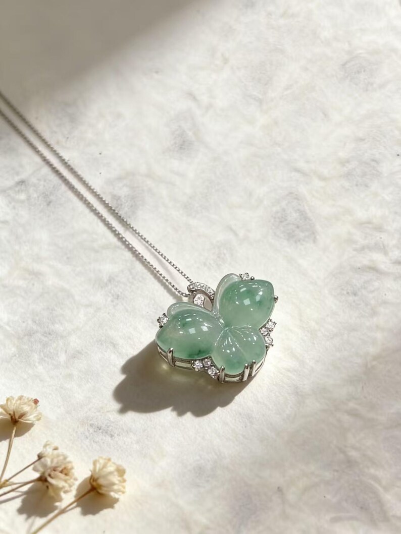 Puede incluir: Un collar de plata con un colgante en forma de mariposa. El colgante est&aacute; hecho de jade verde transl&uacute;cido y est&aacute; adornado con peque&ntilde;as piedras preciosas transparentes. El collar se muestra sobre una superficie blanca texturizada.