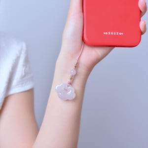 Könnte beinhalten: Eine rote Handyhülle mit dem Text "2022" wird in einer Hand gehalten. Ein rosa Band mit Perlen und einem wolkenförmigen Anhänger hängt am Telefon. Der Anhänger hat eine hellrosa Farbe.
