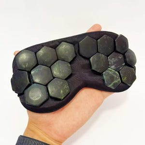 May include: A black eye mask featuring numerous dark green, hexagonal jade stones. The stones are arranged across the mask's surface. The mask is held in a hand against a white background.