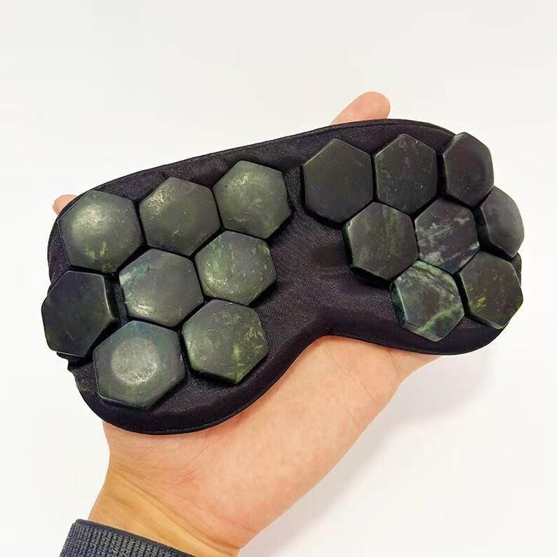 May include: A black eye mask featuring numerous dark green, hexagonal jade stones. The stones are arranged across the mask's surface. The mask is held in a hand against a white background.