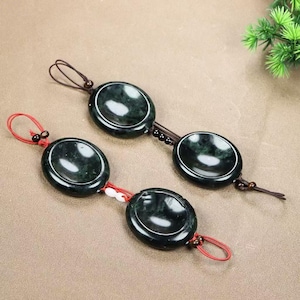 May include: Two dark green jade pendants, each composed of three oval-shaped stones. The stones are connected by brown and red cords. The pendants have a polished, glossy finish, and are displayed on a neutral-toned surface.