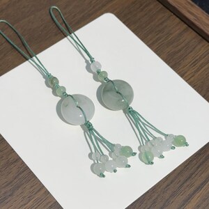 May include: Two decorative charms featuring circular pale green jade-like stone discs suspended on sage green braided cords. Each charm includes smaller translucent white and green beads arranged along the cord and at the base of the tassel.