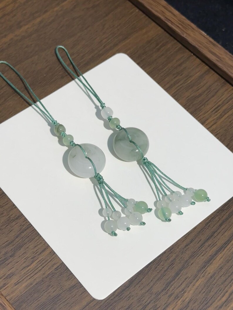 May include: Two decorative charms featuring circular pale green jade-like stone discs suspended on sage green braided cords. Each charm includes smaller translucent white and green beads arranged along the cord and at the base of the tassel.