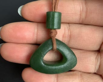 May include: A dark green jade pendant with a triangular shape and a hole in the center. The pendant is suspended from a brown cord, with a cylindrical jade bead above it. The pendant is held in a person's hand.