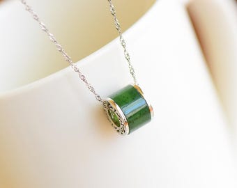 Puede incluir: Un collar de plata con un colgante cilíndrico de jade verde. El jade está engastado en un marco de plata decorativo, y la cadena tiene un diseño delicado y retorcido. El colgante mide aproximadamente 2 cm de largo.