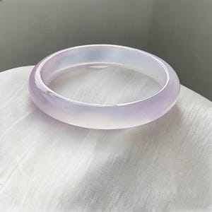 Puede incluir: Un brazalete de jade translúcido de color lavanda. La superficie pulida refleja la luz, mostrando las variaciones naturales de color. El brazalete tiene forma circular y descansa sobre una superficie blanca texturizada.