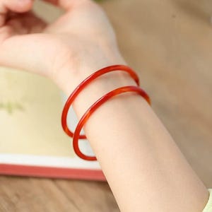 Puede incluir: Dos brazaletes translúcidos de color rojo-naranja adornan una muñeca. Las pulseras son finas y circulares, con un acabado brillante. La imagen es un primer plano, que destaca el color y la forma de las joyas.