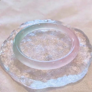 Può includere: Un braccialetto bangle di giada traslucida con una sfumatura di colore, che passa dal verde pallido al rosa tenue. Il braccialetto poggia su una lastra di vetro trasparente, strutturata e di forma irregolare, creando un'estetica delicata ed elegante.