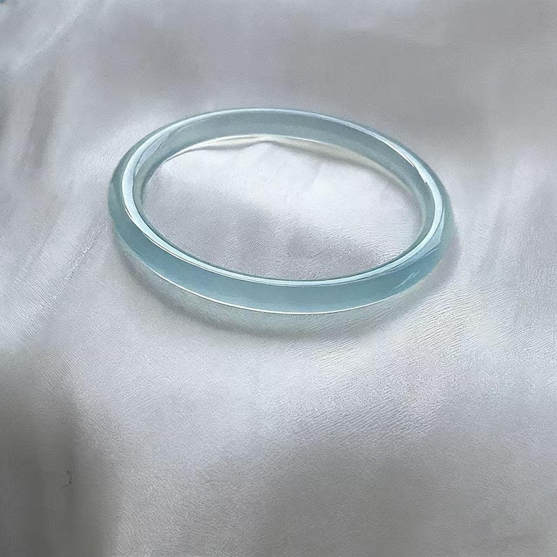 May include: A light blue, translucent bangle bracelet. The bracelet is a solid, circular band with a smooth, polished surface. The bangle is resting on a white, silky fabric.