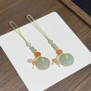 May include: Two decorative charms featuring pale green jade-like circular rings, accented with smaller orange and translucent white beads. Each charm is suspended from a thin, light yellow cord loop, creating a symmetrical pair of ornaments.