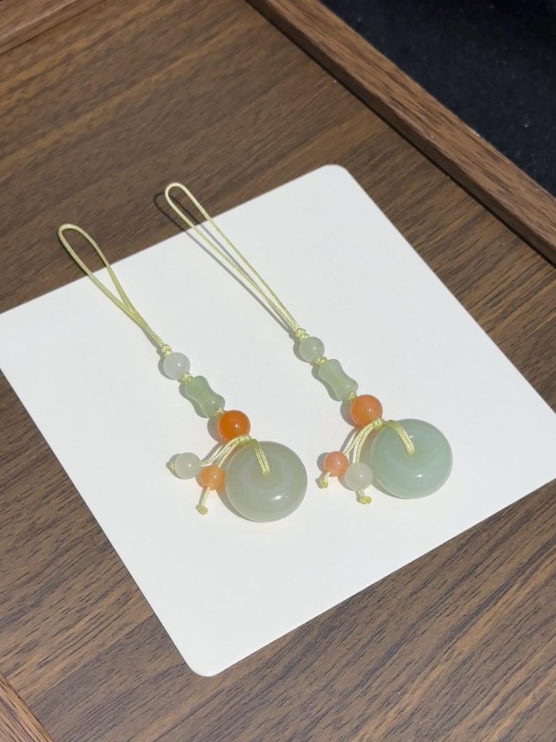 May include: Two decorative charms featuring pale green jade-like circular rings, accented with smaller orange and translucent white beads. Each charm is suspended from a thin, light yellow cord loop, creating a symmetrical pair of ornaments.