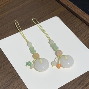May include: Two matching phone charms featuring a central white jade-like donut bead, accented with smaller green, orange, and translucent beads. Each charm is suspended by a thin yellow cord, creating a delicate and symmetrical decorative accessory.