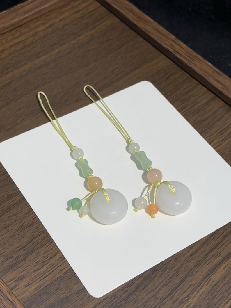 May include: Two matching phone charms featuring a central white jade-like donut bead, accented with smaller green, orange, and translucent beads. Each charm is suspended by a thin yellow cord, creating a delicate and symmetrical decorative accessory.